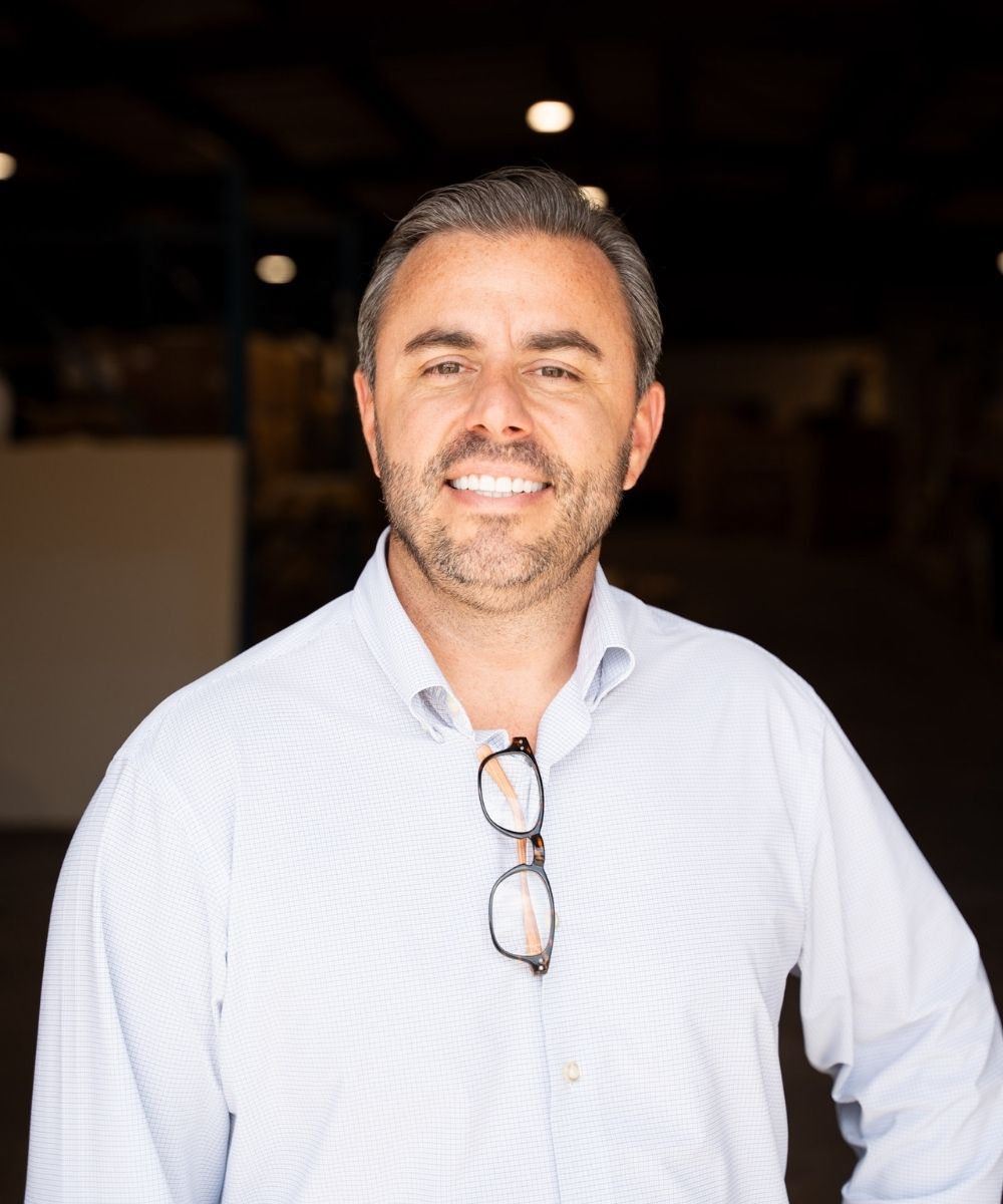 A man with short dark hair and a beard smiles at the camera. He is wearing a light blue button-down shirt with eyeglasses hanging from the collar, standing in a dimly lit indoor space.