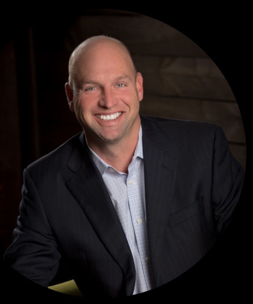 A smiling man with a bald head wearing a dark suit jacket over a light-colored dress shirt poses in front of a dark, blurred background.